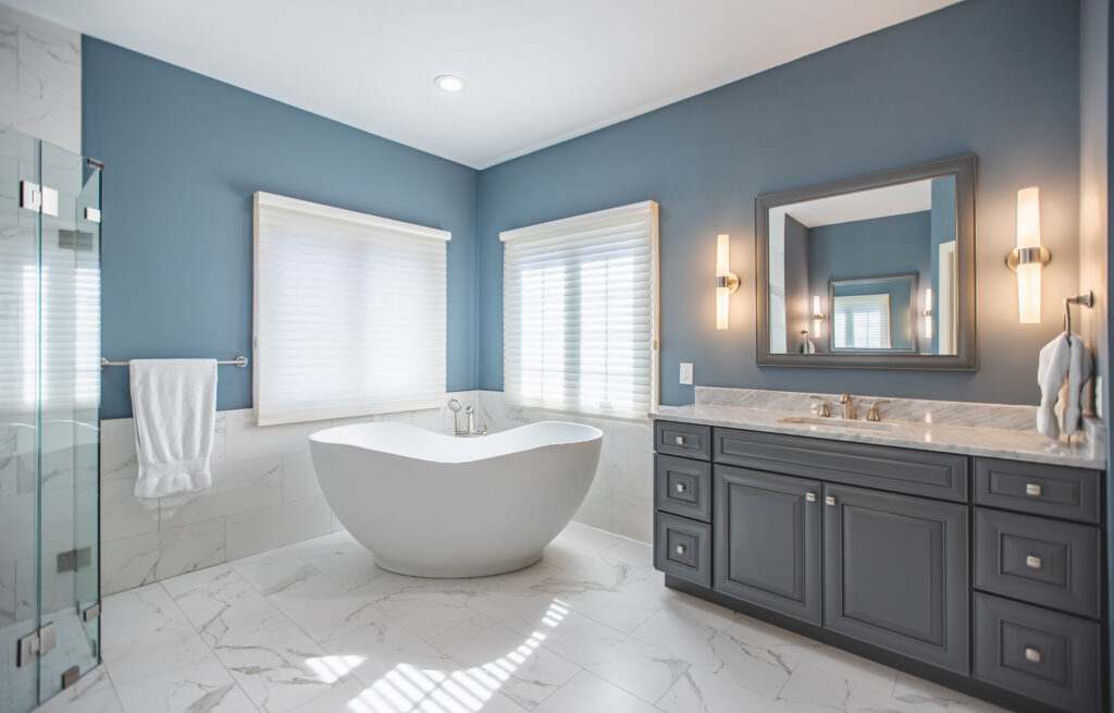 Bathroom remodeling in Sterling VA with freestanding soaking tub, blue accent walls, gray vanity with marble countertop, gold sconces, and frameless glass shower