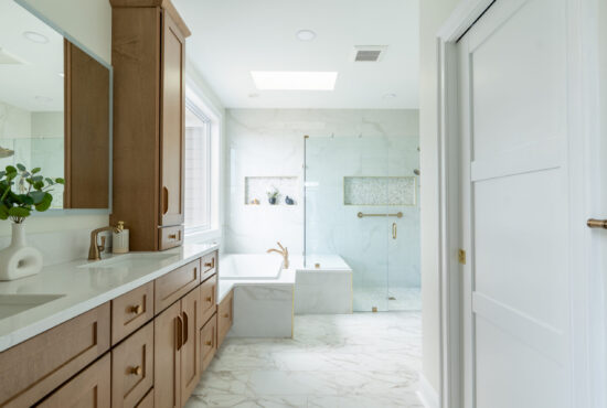 Bathroom remodeling in Sterling VA featuring a wood vanity with gold fixtures, frameless glass shower, soaking tub, recessed niche, and skylight